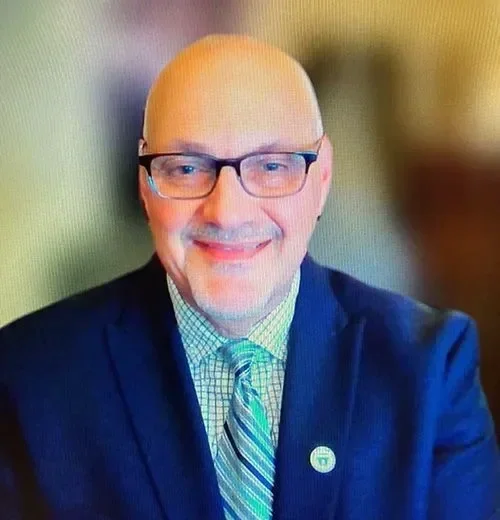 A bald man wearing glasses, a checkered shirt, a striped tie, and a suit jacket, smiling at the camera.