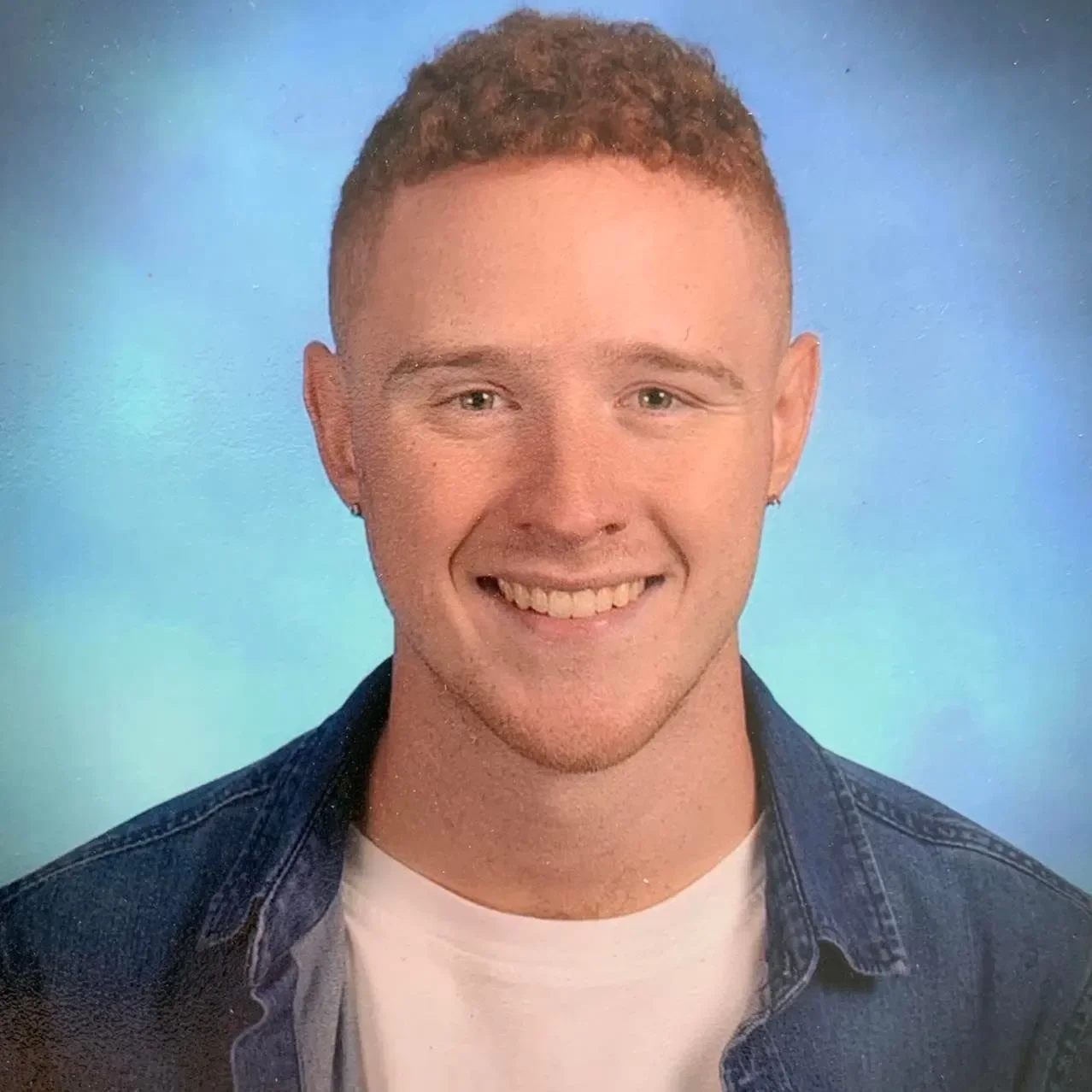 A young man with short, curly red hair, blue eyes, and a bright smile wearing a denim jacket over a white t-shirt, posing against a blue background.