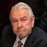 Portrait of an elderly man with white hair and a beard, wearing a dark suit and a patterned tie.