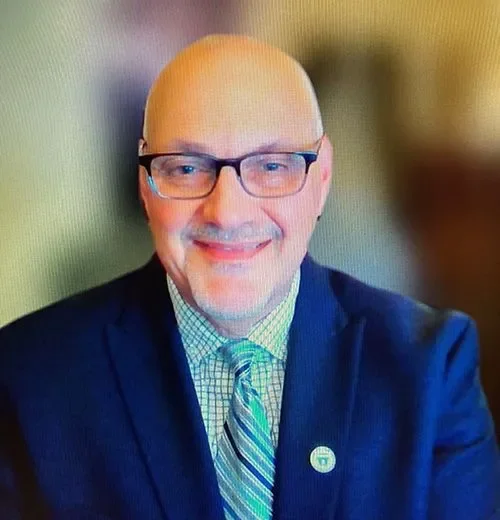 A smiling man with glasses, wearing a suit and tie, in front of a blurred background.