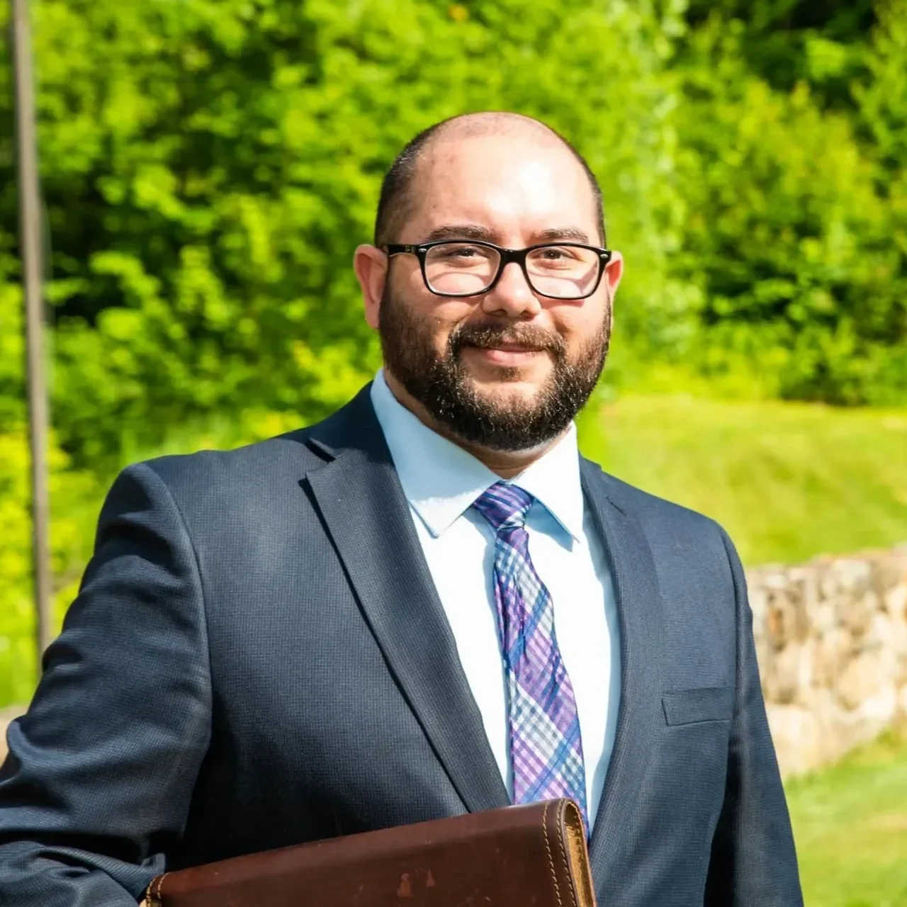 A man in a dark suit, white shirt, and plaid tie standing outdoors with green trees in the background. He has glasses, a beard, and is holding a brown leather folder.