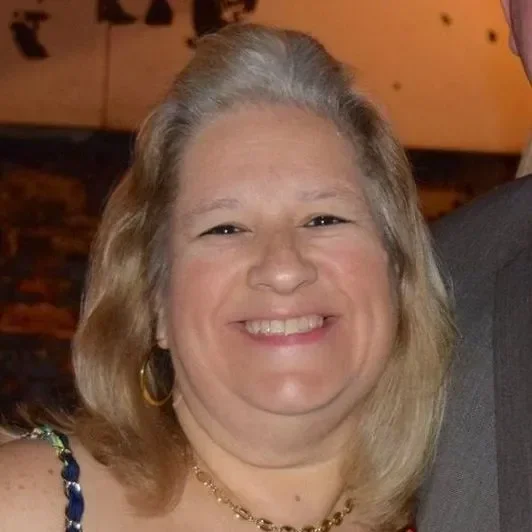 A smiling woman with shoulder-length blonde hair, wearing gold hoop earrings and a necklace, in an indoor setting with warm lighting.