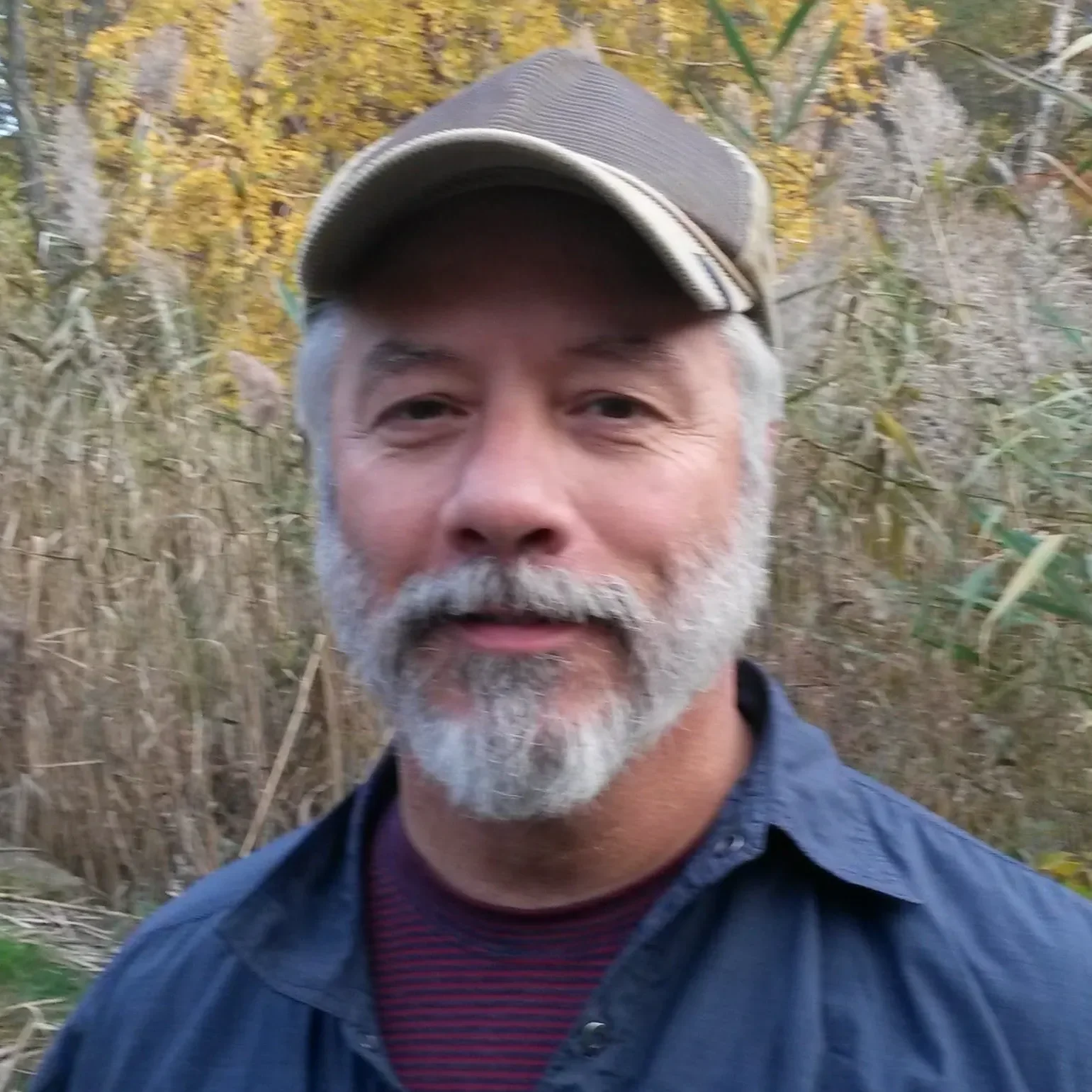 A middle-aged man with a gray beard and mustache, wearing a cap and a dark shirt, standing outdoors in a natural setting with tall grasses and trees in the background.