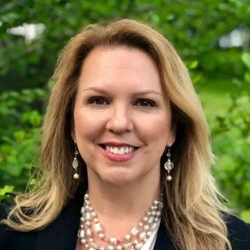Portrait of a woman with blonde hair smiling, wearing earrings and a pearl necklace, outdoors with green foliage in the background.