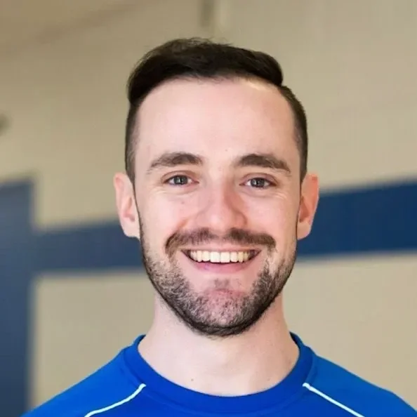 A smiling man with short dark hair, a beard, and blue eyes, wearing a blue athletic shirt, standing indoors with a blurred background.