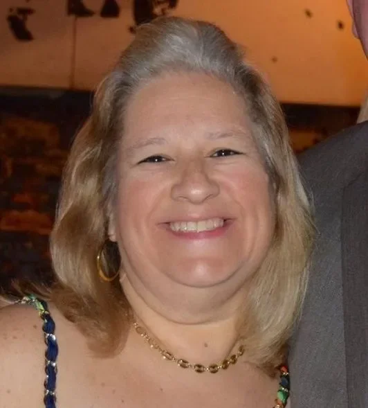 A woman with blonde hair smiling, wearing gold hoop earrings, a gold chain necklace, and a colorful dress, standing indoors.