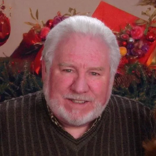 An elderly man with white hair and a beard smiling, in front of a Christmas-themed background with red and gold decorations.