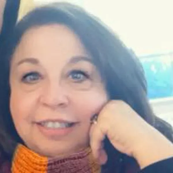 Close-up of a woman with dark hair and blue eyes smiling, resting her face on her hand, with a blurred background.