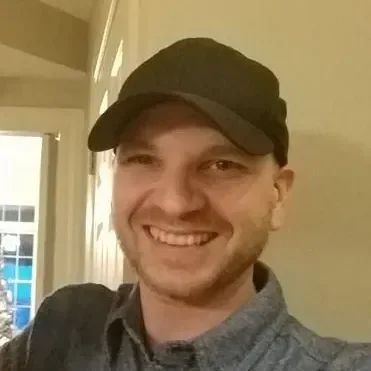 A smiling man wearing a black cap and a gray button-up shirt, indoors with a window and a room behind him.
