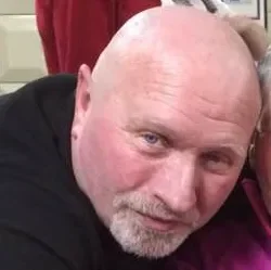 Close-up of an older man with a bald head and gray facial hair, wearing a black shirt, looking at the camera.