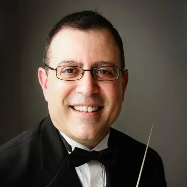 Portrait of a man with glasses, smiling, wearing a tuxedo and bowtie, holding a conductor's baton.