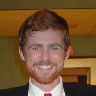 A smiling man with a beard and mustache, wearing a dark suit and red tie, indoors with a yellow ceiling and green walls.