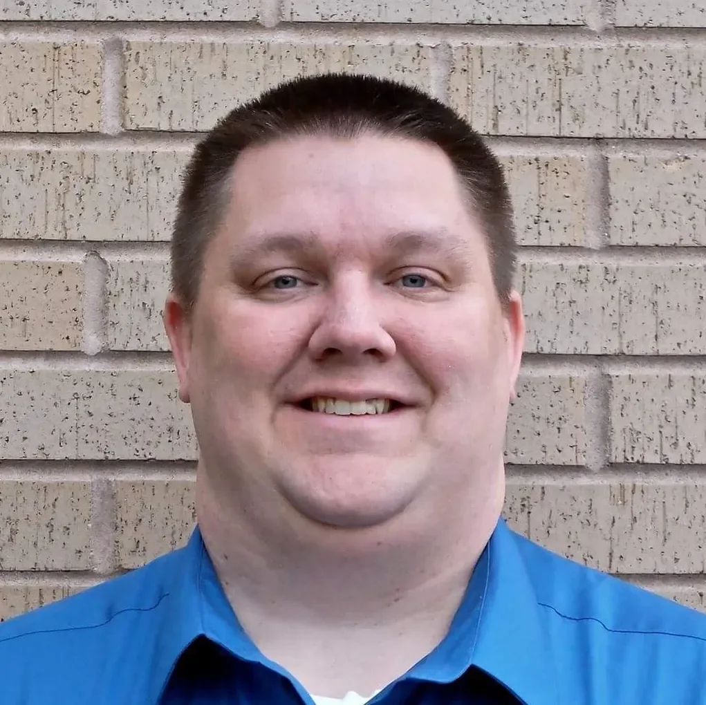 A man with short dark hair, light skin, and blue eyes smiling in front of a beige brick wall. He is wearing a bright blue shirt.
