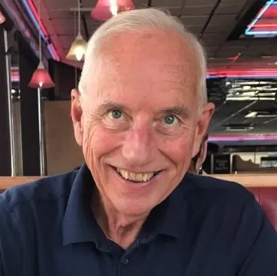 A smiling older man with short white hair wearing a dark blue shirt in a restaurant or bar with colorful pendant lights and neon signs in the background.