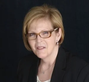 A woman with short blonde hair wearing glasses and a black blazer, making a stern or confused facial expression against a dark background.