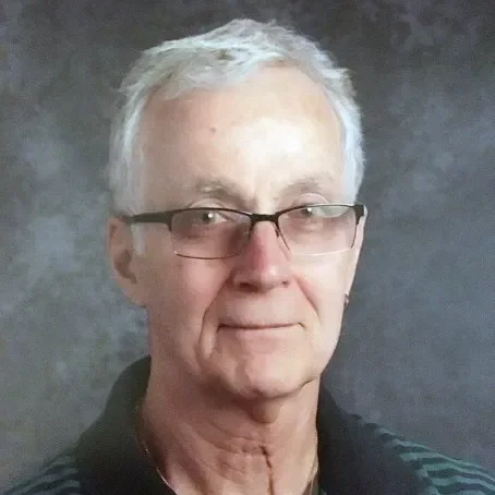 Close-up of an older man with white hair, glasses, and a dark striped polo shirt, standing against a dark background.