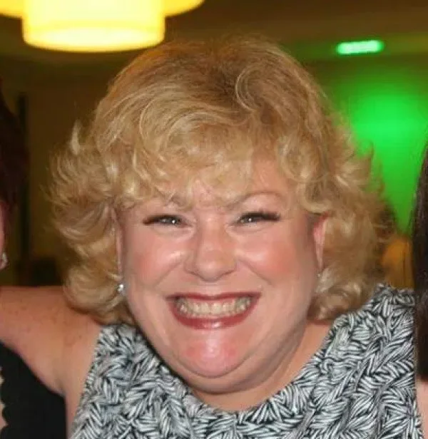 A woman with curly blonde hair smiling widely at a social event, wearing earrings and a patterned top, with colorful ambient lighting in the background.
