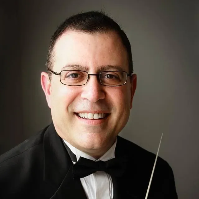A man in a tuxedo with glasses, smiling, holding a conductor's baton.