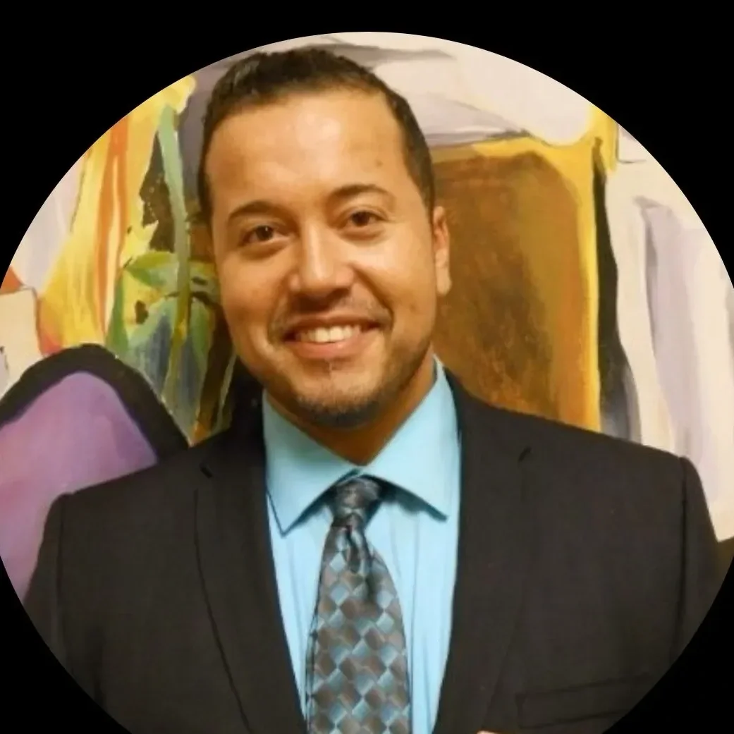 A man in a suit with a patterned tie and light blue shirt smiling, standing in front of a colorful abstract painting.