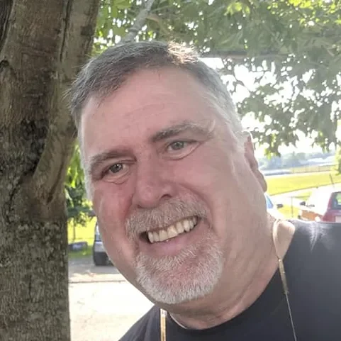 A smiling man with gray hair and a beard outdoors near a tree, with cars and trees in the background.