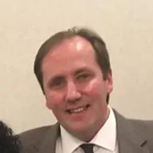 A smiling man with dark hair, wearing a suit and tie, in front of a plain wall.