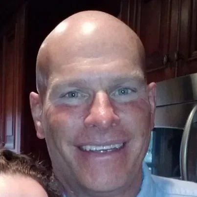 Close-up of a smiling bald man with light skin, blue eyes, and a light blue shirt in a kitchen with dark wooden cabinets.