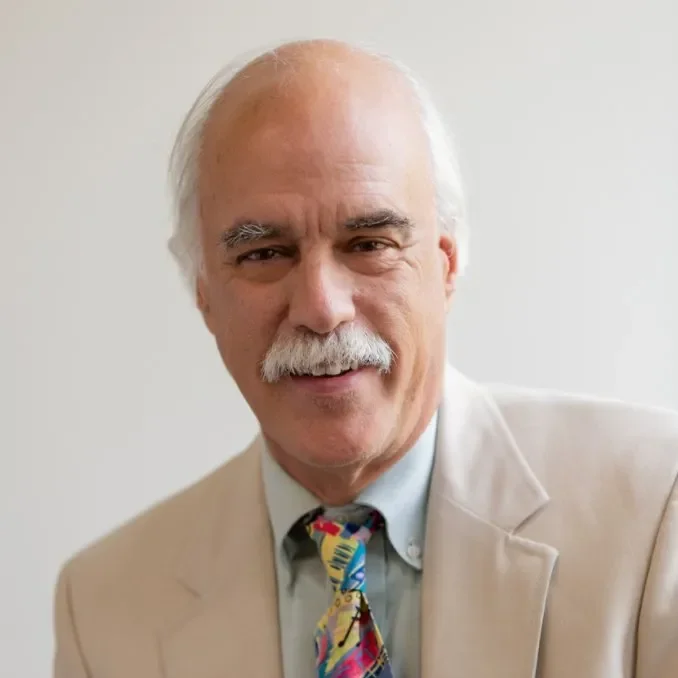 Portrait of an elderly man with white hair and a mustache, wearing a beige suit jacket, a light blue shirt, and a colorful tie, smiling in front of a light-colored background.
