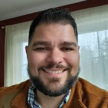 A man with dark hair and a beard smiling inside a room with a window and curtains in the background.