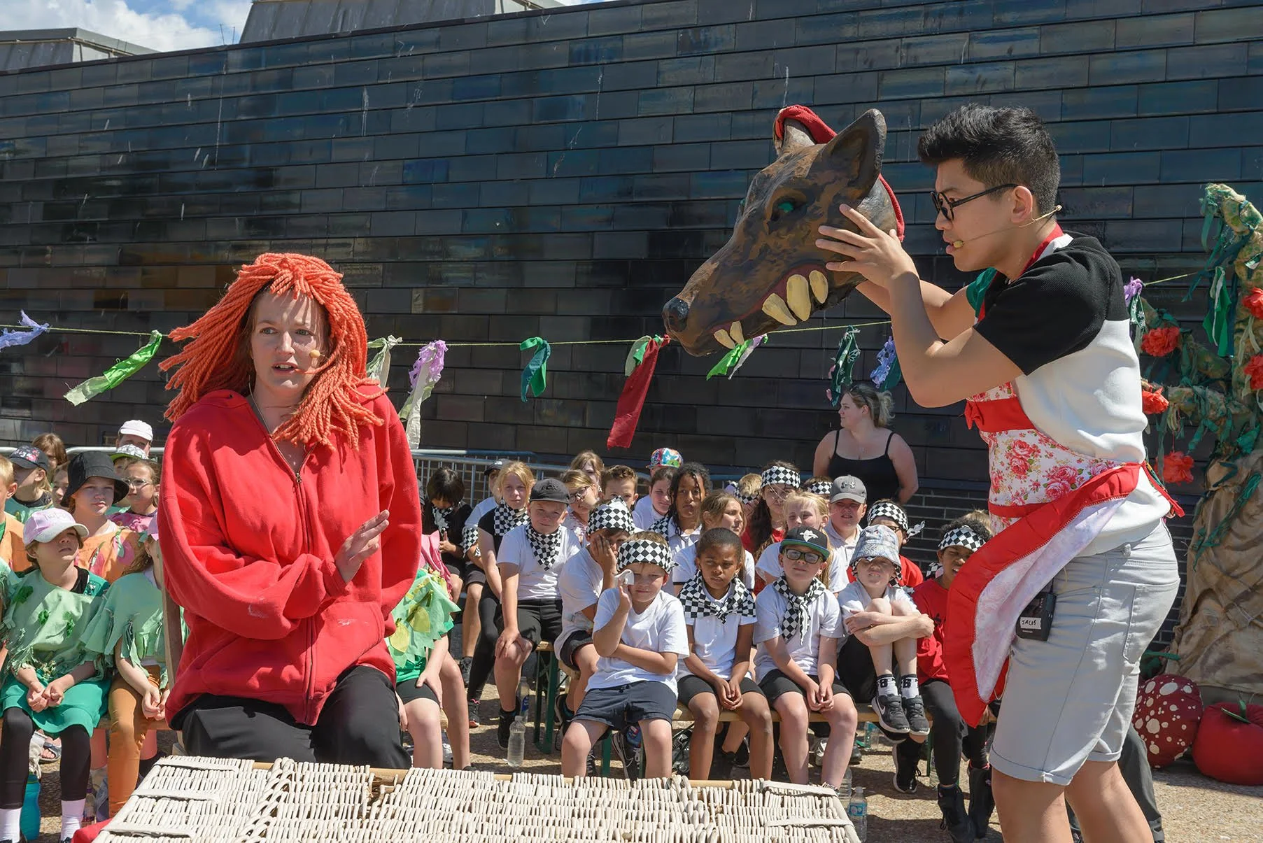 A puppet show with a person wearing a red hoodie and a person holding a wolf puppet, with a group of children seated watching outdoors.