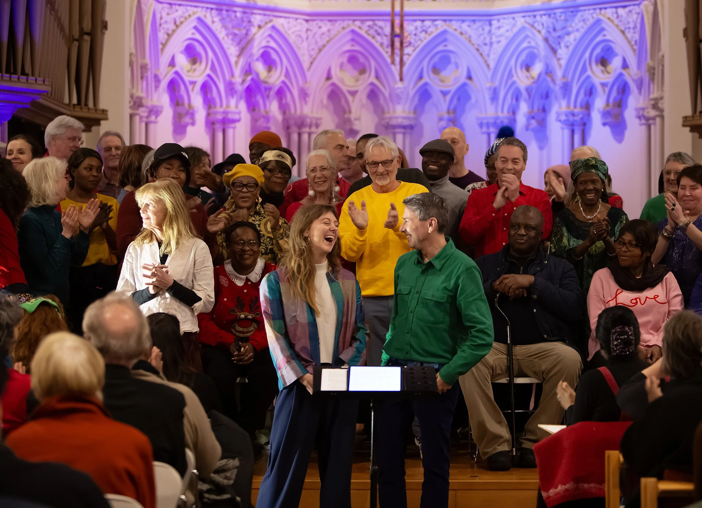 A diverse group of people gathered in a church or concert hall. At the center, a woman and a man are standing, smiling, and looking at each other. The woman is wearing a colorful, striped jacket, and the man is wearing a bright green shirt. Behind th