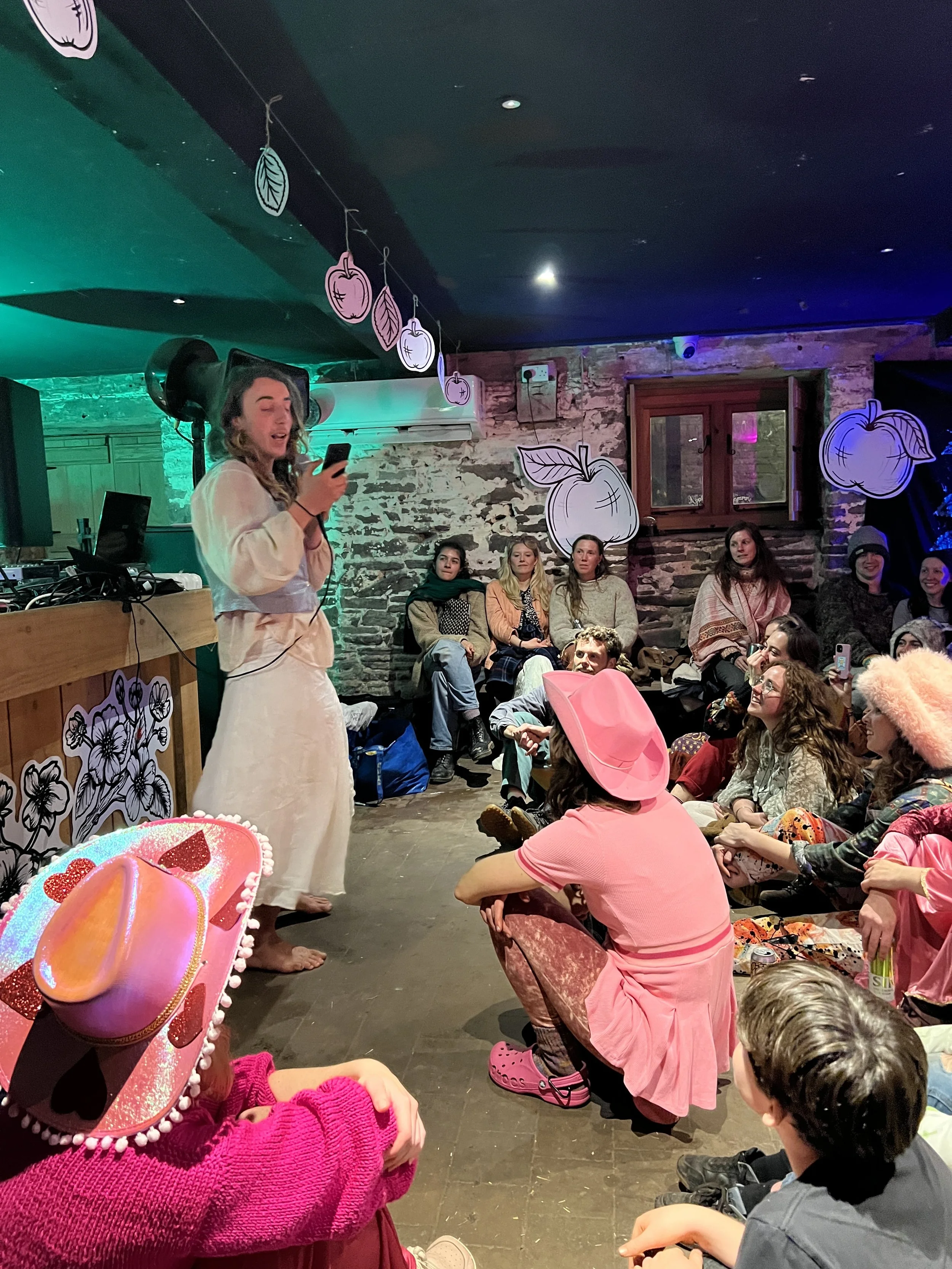 A woman dressed in a white outfit speaking or singing to an audience at a small indoor gathering, surrounded by people wearing colorful costumes, some sitting on the floor and others on chairs, with decorations including paper cutouts of apples and l