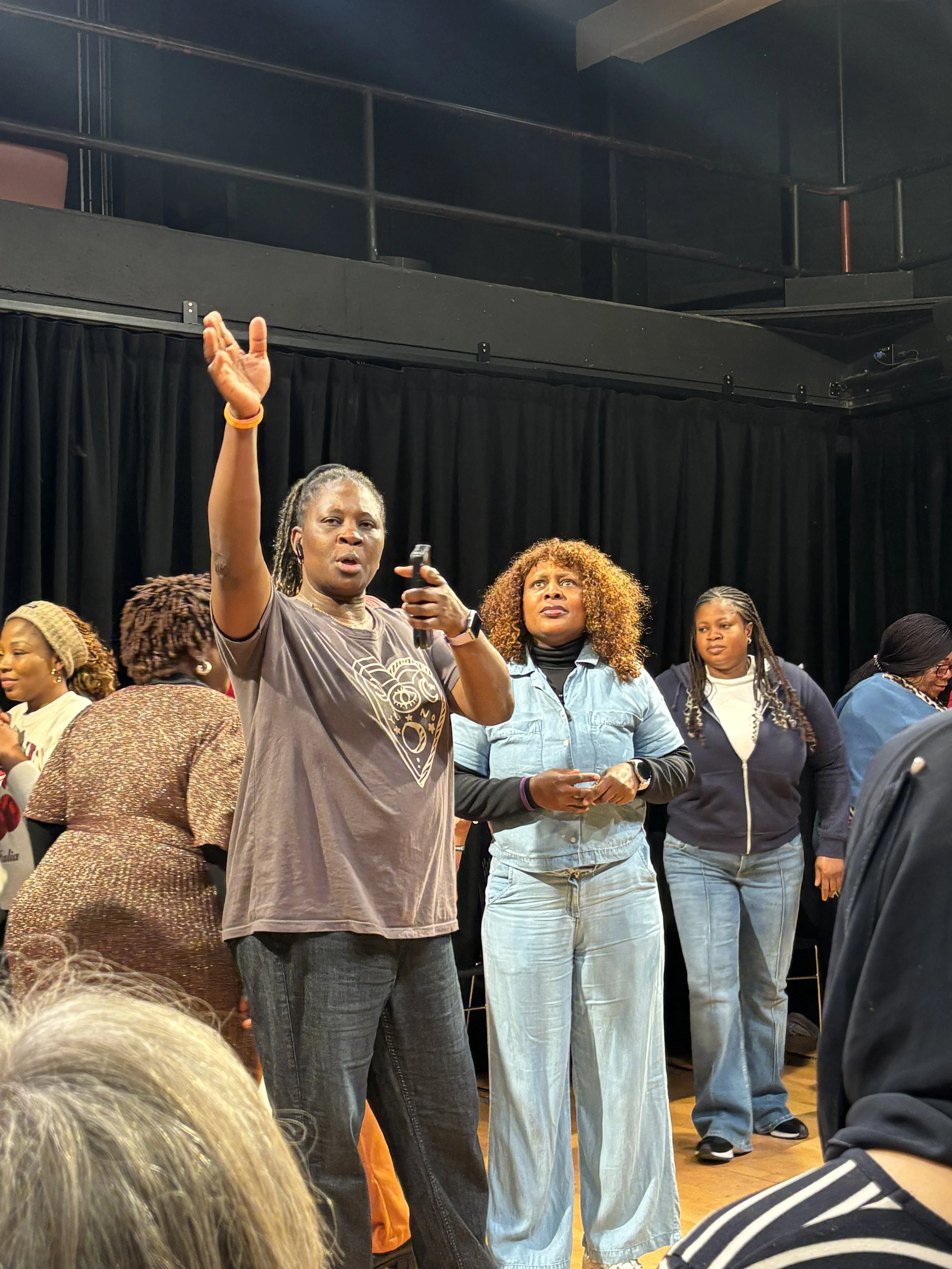 A group of women standing on a stage, one woman speaking and gesturing with her hand, while others listen. The setting appears to be an indoor event or gathering.