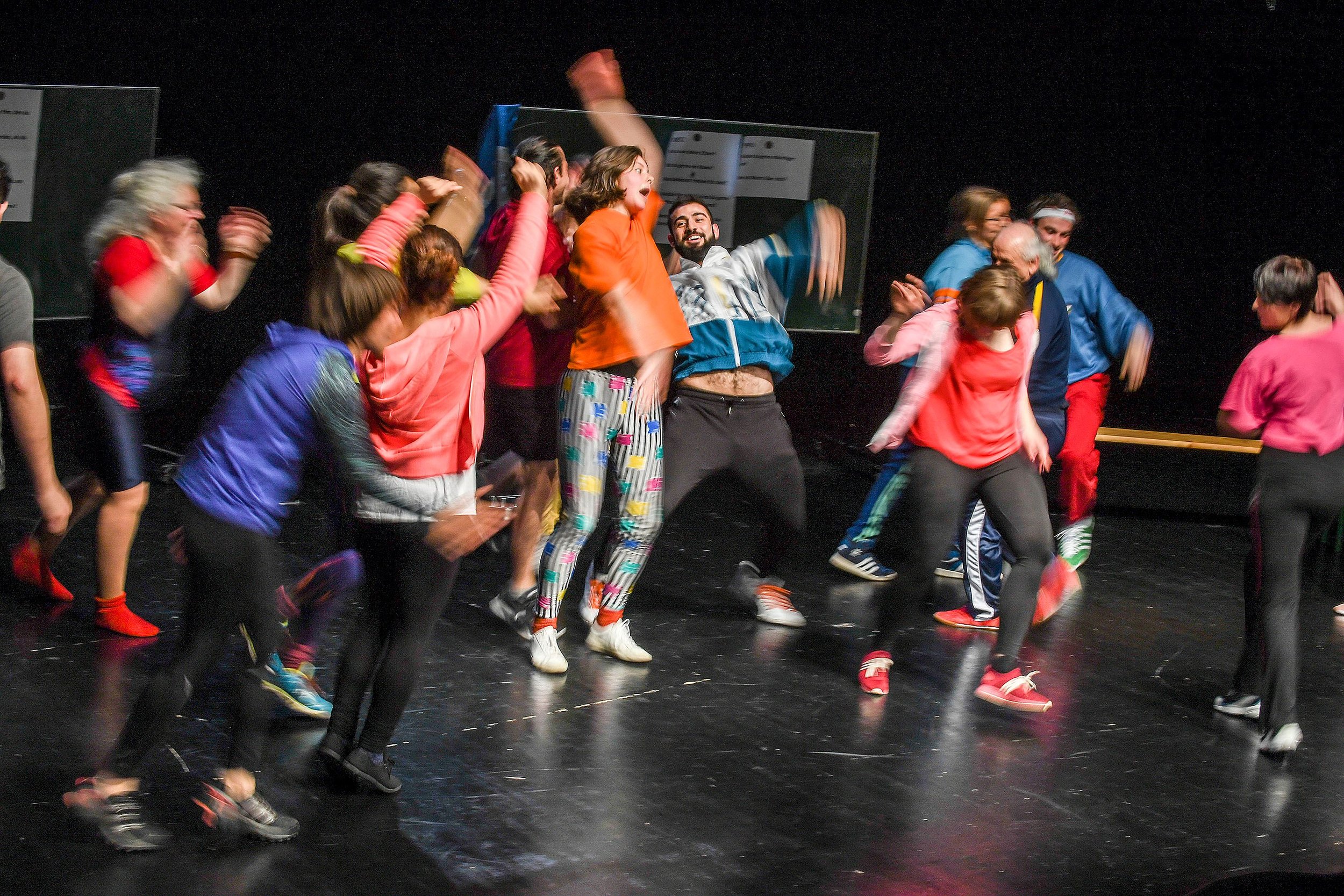 A group of people on stage dancing and celebrating, dressed in colorful athletic clothing, with some wearing glasses and headbands, against a black background.