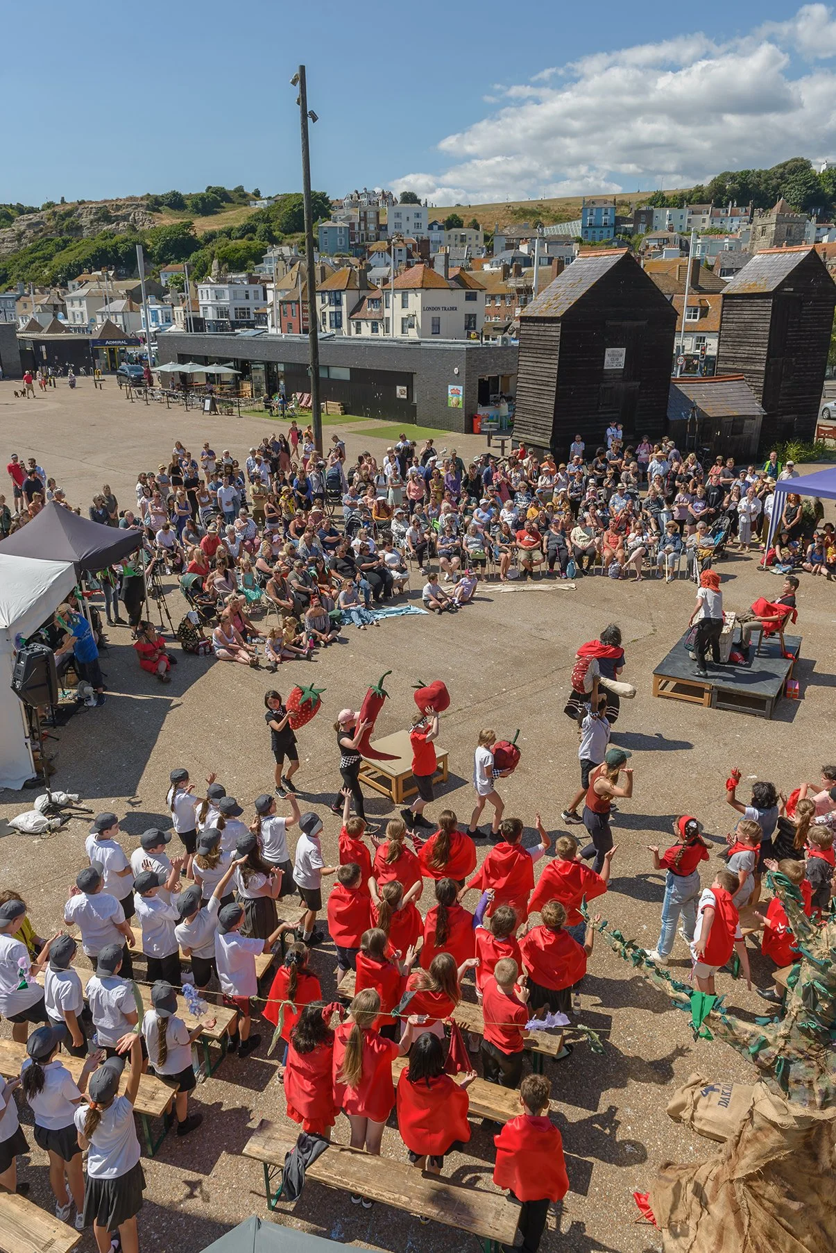 Children and adults participating in a strawberry-themed event outdoors, with some children in white shirts and gray hats, others in red, and a stage where some people are performing or presenting.