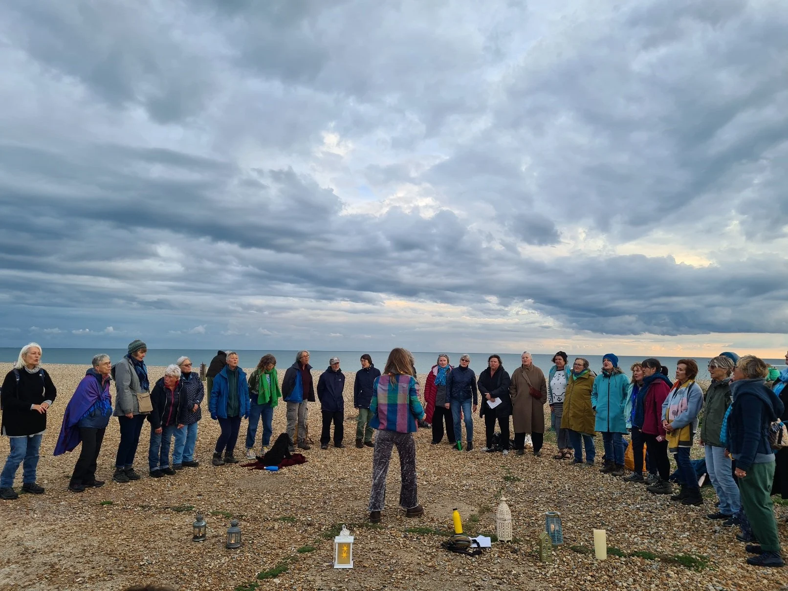 A group of people standing in a circle on a pebble beach, facing inward, with a person in the center. The sky is cloudy, and the ocean is visible in the background. Items such as lanterns, candles, and small decorative objects are placed on the groun