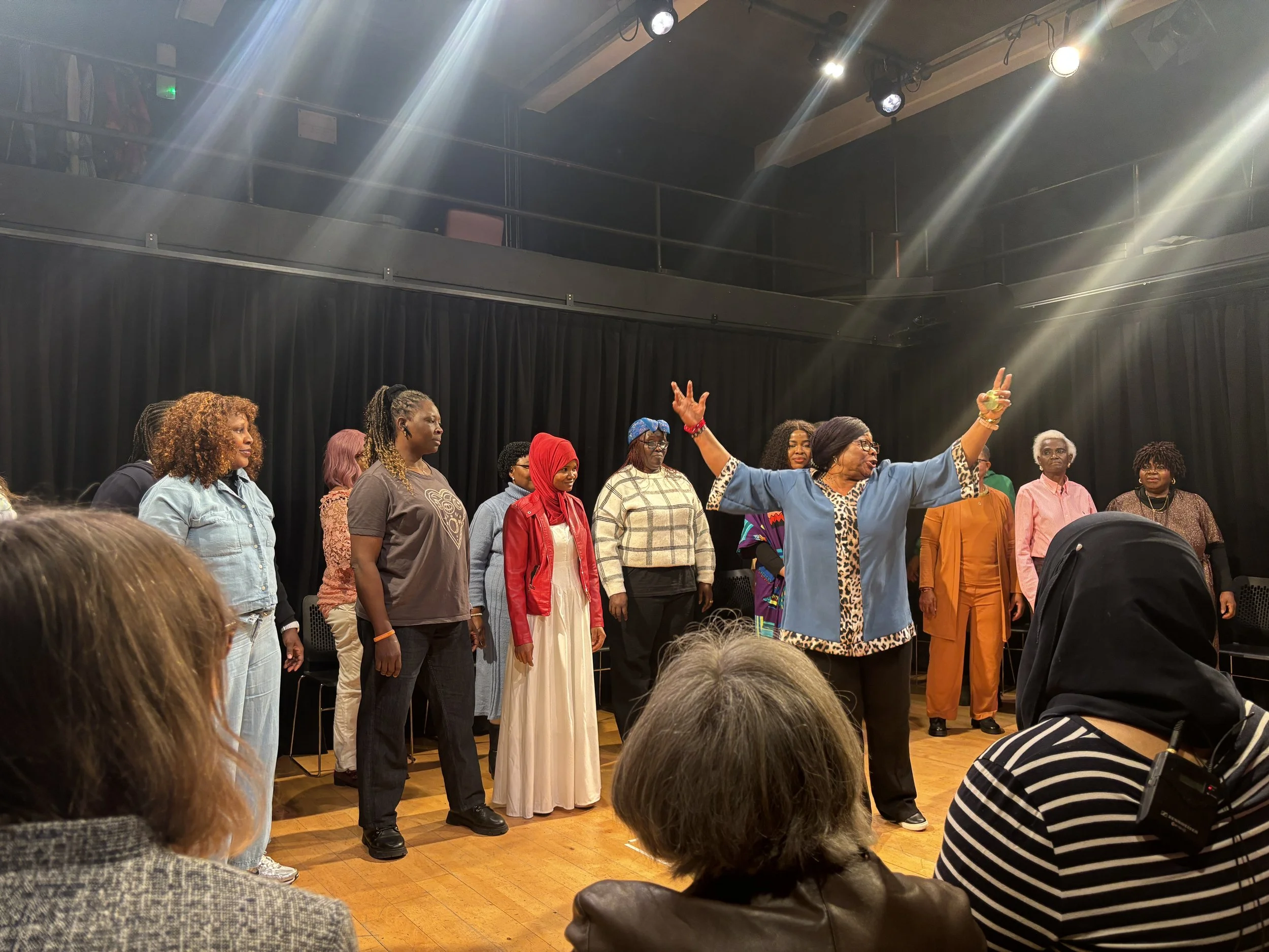 A group of women standing on a stage in a theater, with one woman in front gesturing passionately with arms raised, while others watch. Audience members in the foreground observe the scene.