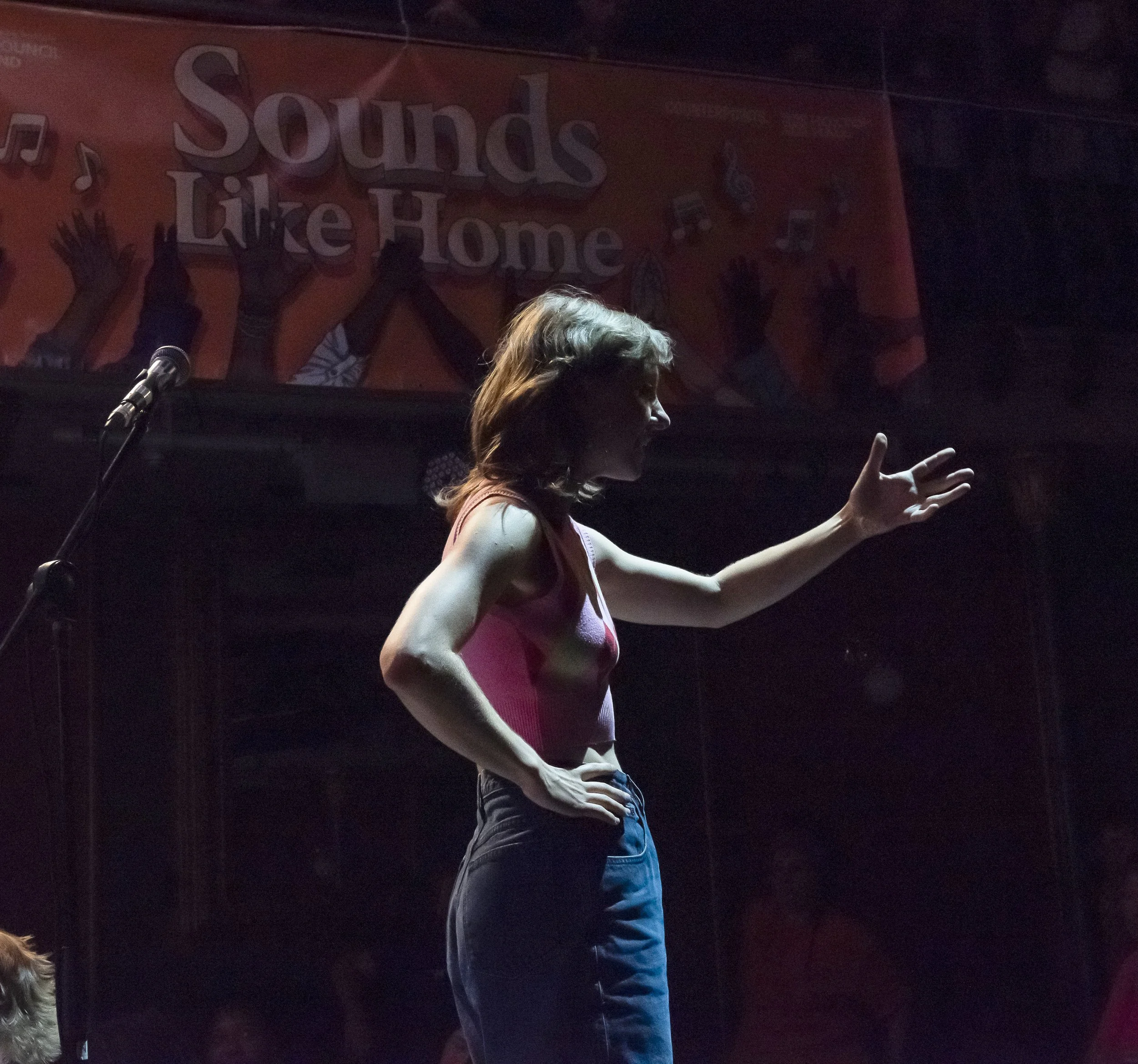 A woman on stage with a microphone in front of her, gesturing with her right hand, under a sign that says 'Sounds like Home' with silhouettes of raised hands and musical notes.