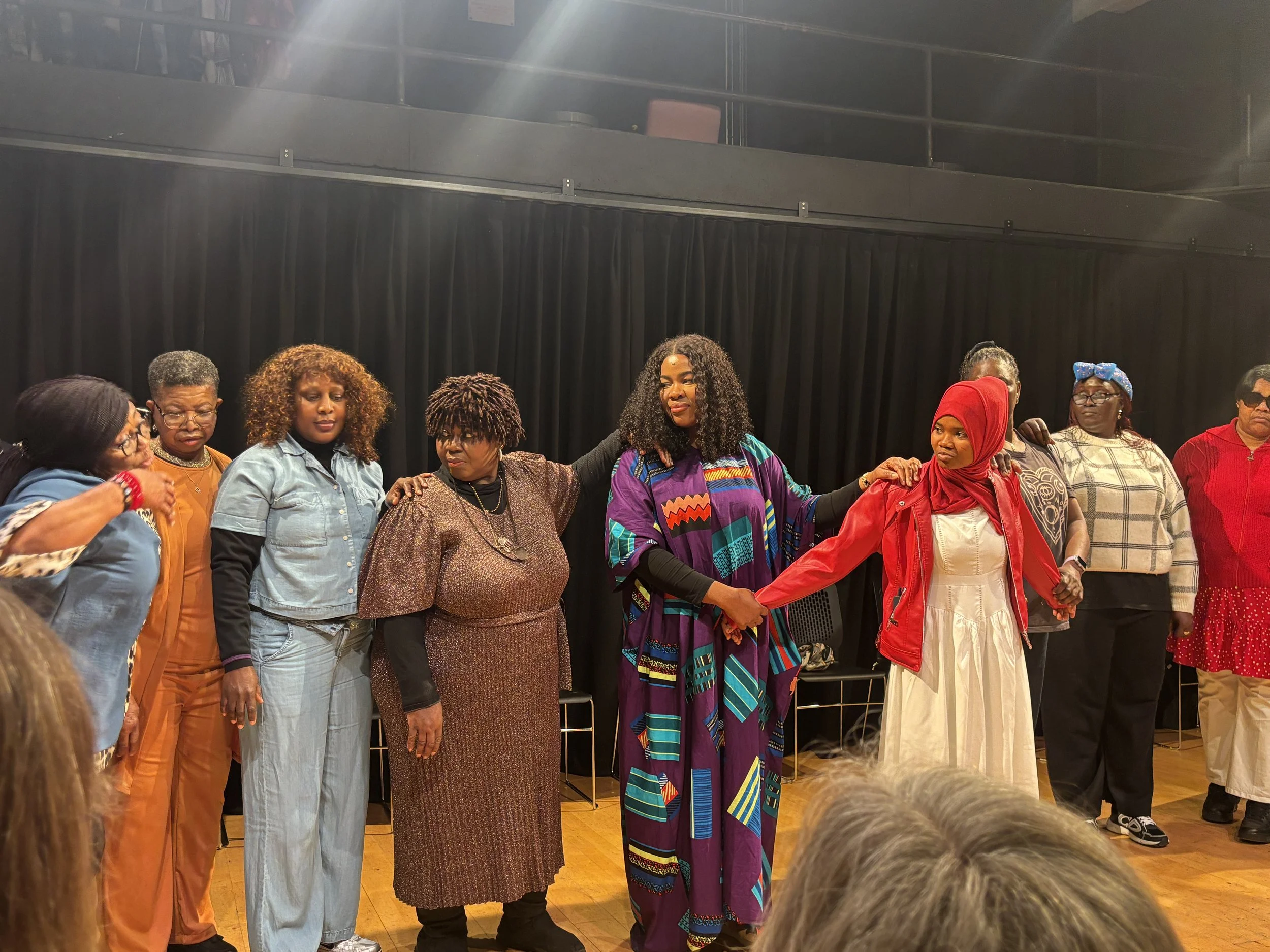 Group of women standing in a line on stage, holding hands, with some placing hands on each other's shoulders, participating in a collective activity or prayer, with a black curtain backdrop.