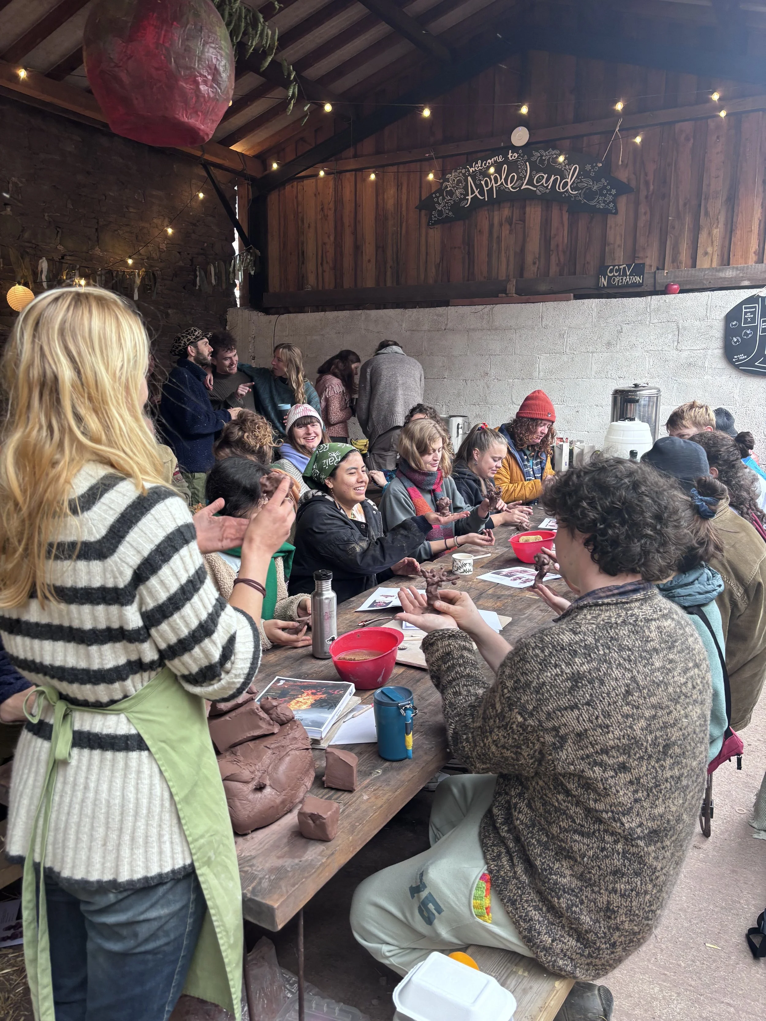 Group of people gathered around a long wooden table indoors, participating in a pottery workshop with clay; some are shaping clay, others are watching and chatting. The room has a rustic ambiance with wooden walls, string lights, and a sign that read