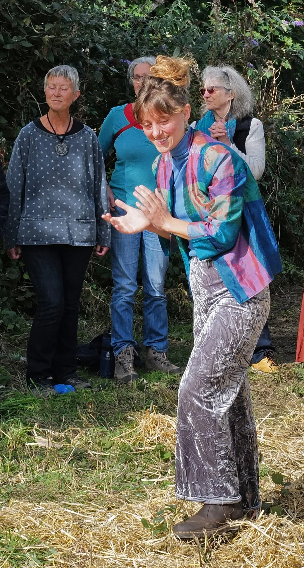 A woman in colorful clothing is clapping and smiling in front of a group of women standing outdoors in front of green shrubs and plants.