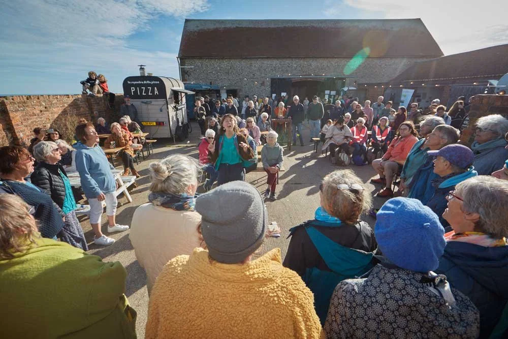 A large group of elderly people gathered outdoors in a courtyard, listening to a woman in a teal jacket speaking. There are picnic benches, a pizza truck, and a stone building in the background, with the sky clear and sunny.