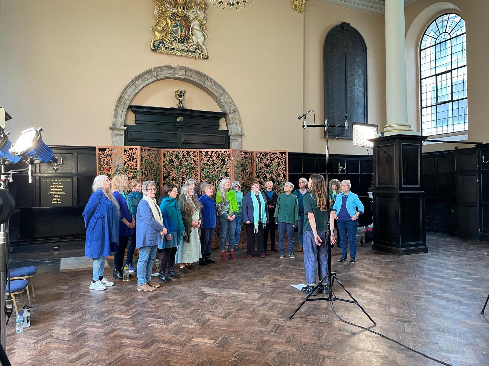 A women's choir practicing in a historic church or hall with tall windows and wooden floors. The choir members are standing in a semi-circle, facing their conductor, as they rehearse. There are professional lights and microphones set up for recording