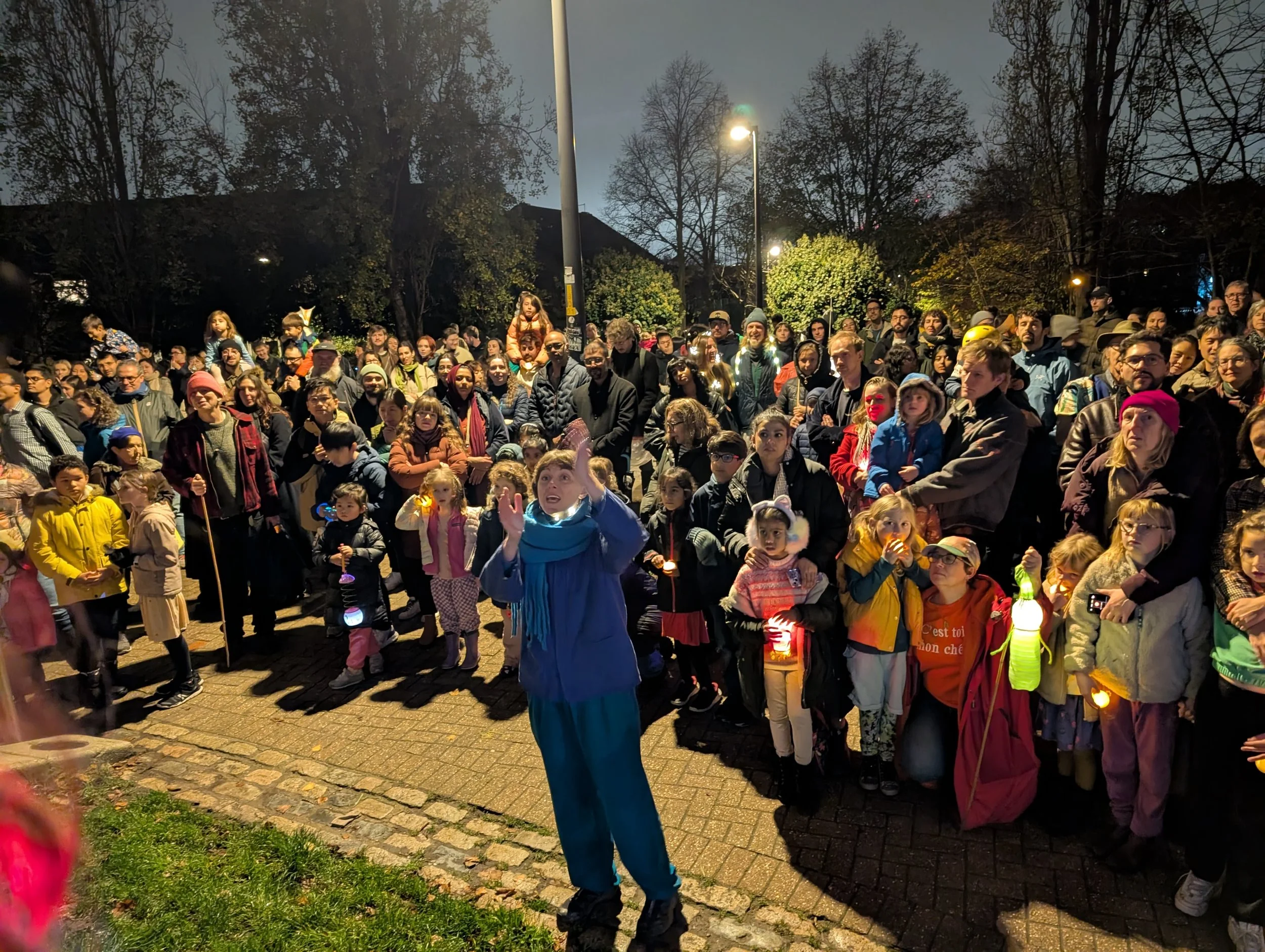 People gathered outdoors at night, some holding lanterns, with a woman in blue jacket speaking to the crowd, surrounded by trees and streetlights.