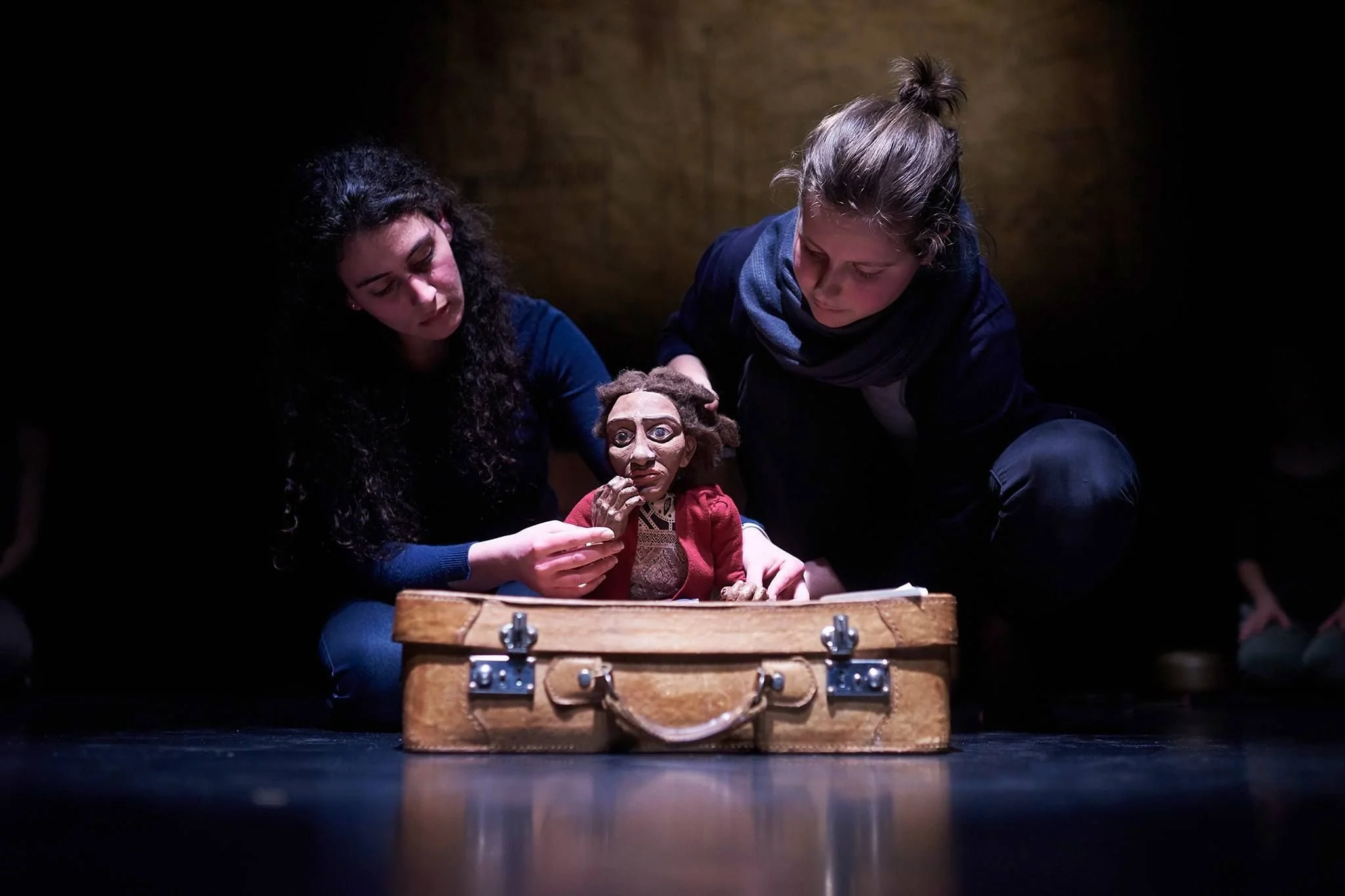 Two women are performing a puppet show with a puppet of a woman, using a wooden case as a stage. They are kneeling on the floor in a dimly lit setting.