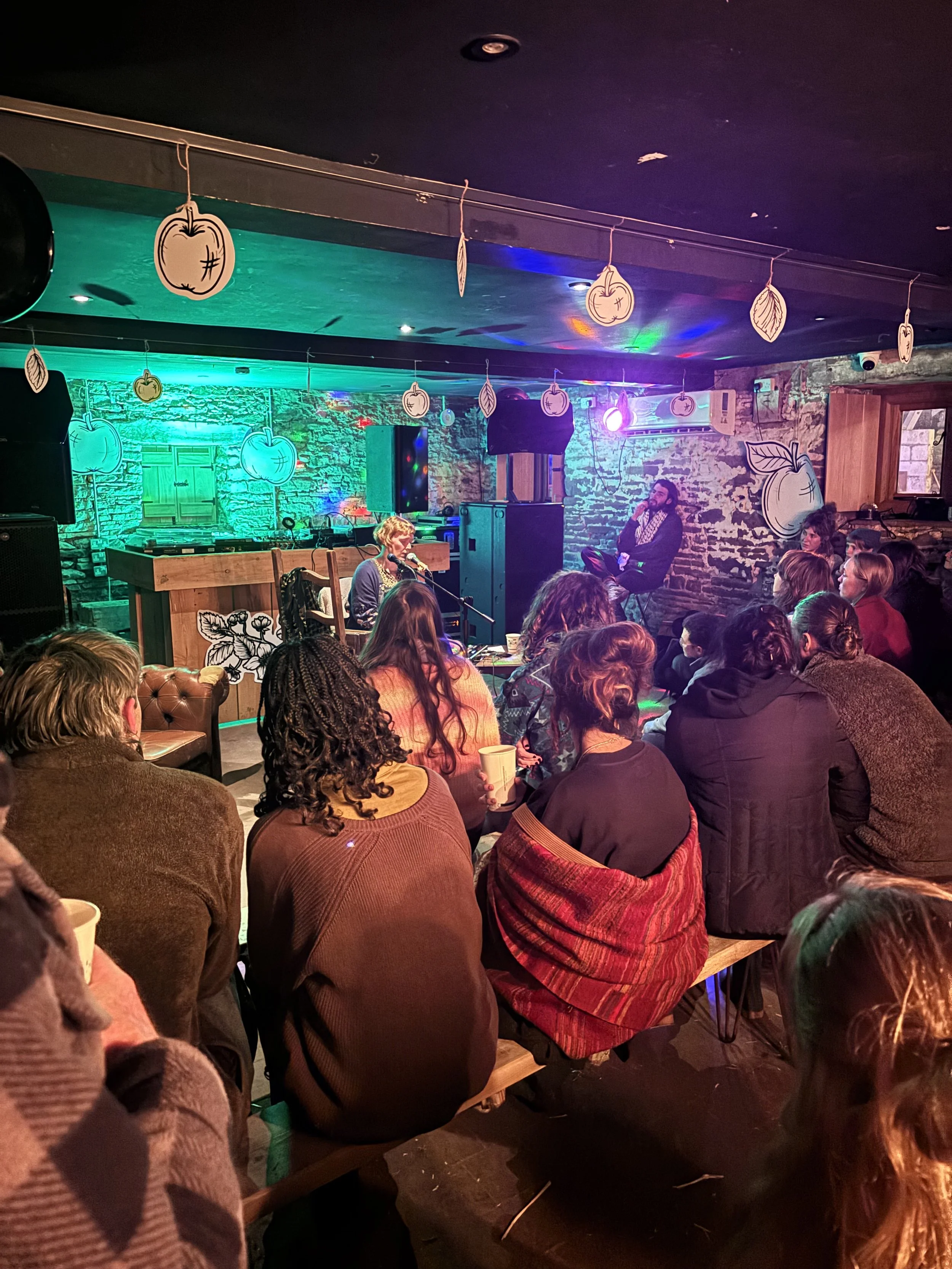 People seated and watching a performance with a woman singing and a man sitting in the background on a stage decorated with hanging drawings of apples and leaves, in a cozy, rustic venue with exposed brick walls.