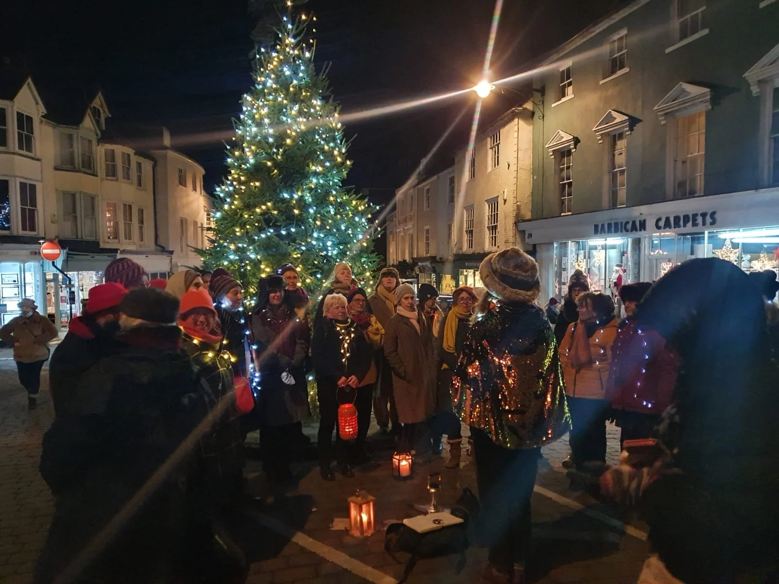 People gathered around a lit Christmas tree on a city street at night, some holding candles or lanterns, with stores and buildings in the background.