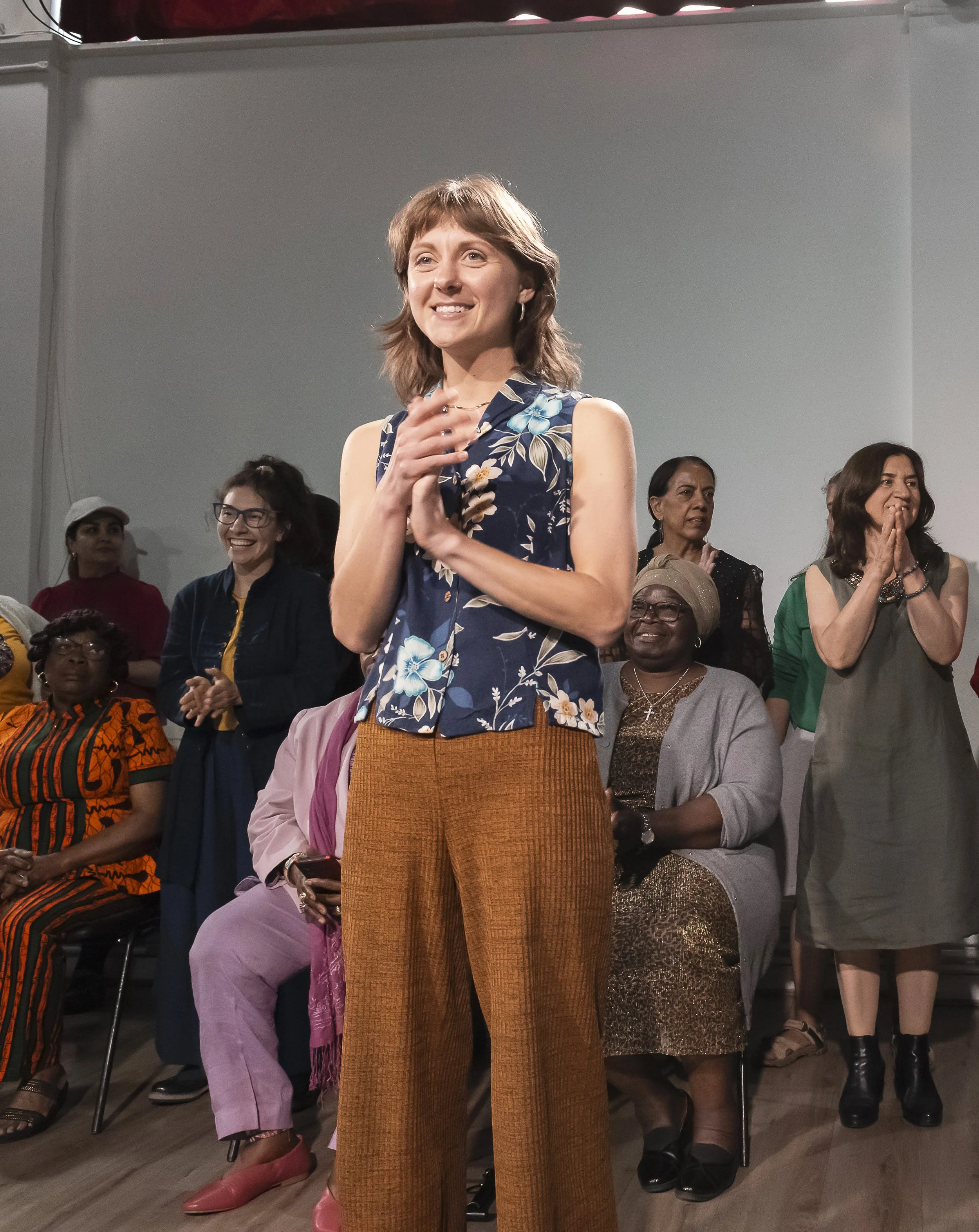 A woman is standing and smiling at an event with several women behind her, some sitting and some standing, in a room with plain walls.