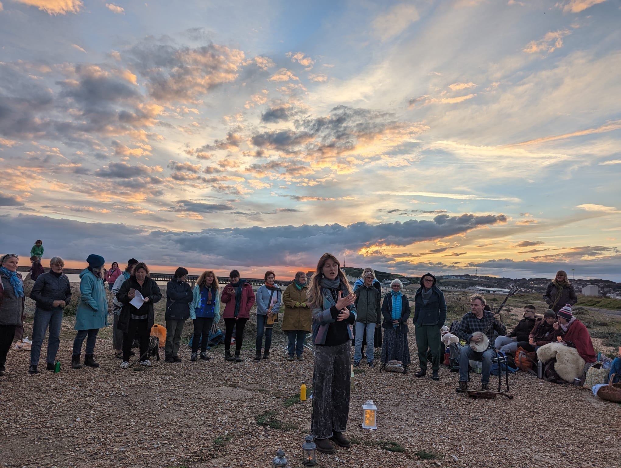 Group of people gathered outdoors during sunset, some sitting and some standing, with a woman speaking in front, holding papers, a few musicians playing instruments nearby, on a gravel area with hills and a cloudy sky in the background.