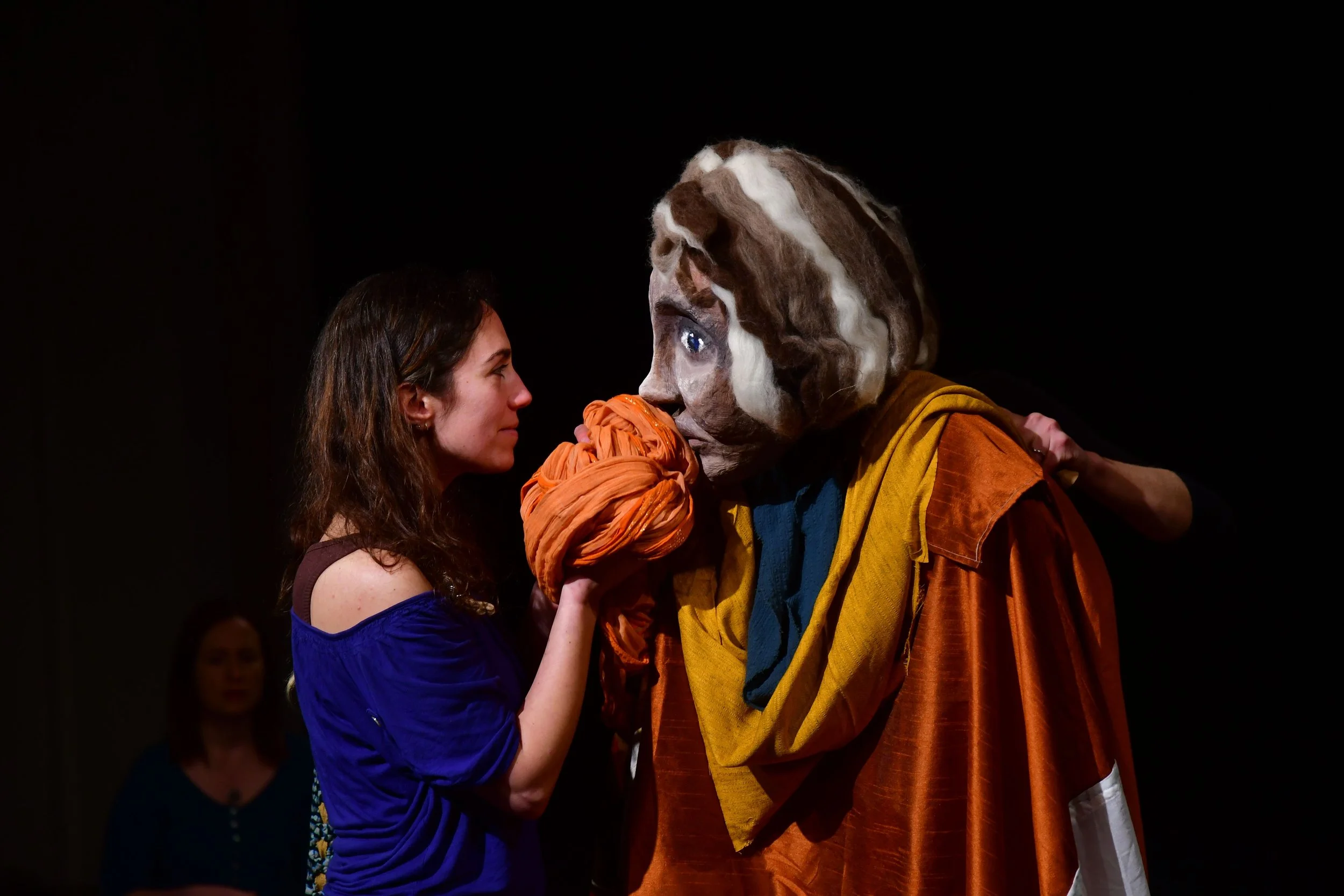 A woman with brown hair in a blue dress facing a person in a large, theatrical mask and costume with a giant beard and colorful robes during a stage performance.