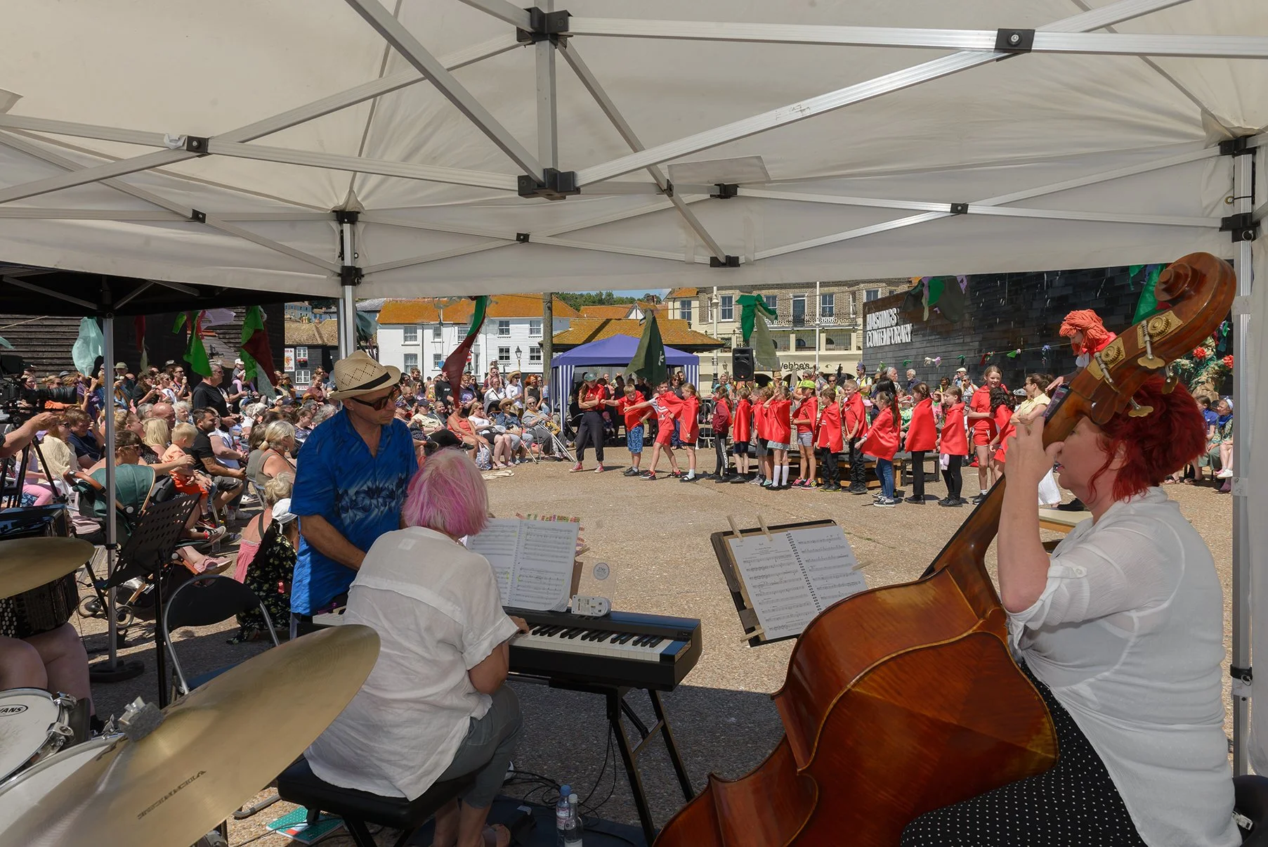 A musical performance taking place at an outdoor festival with a children's choir and an audience seated under a white tent. Musicians playing keyboard, double bass, and drums are visible in the foreground. The children are dressed in red outfits, performing in the background.